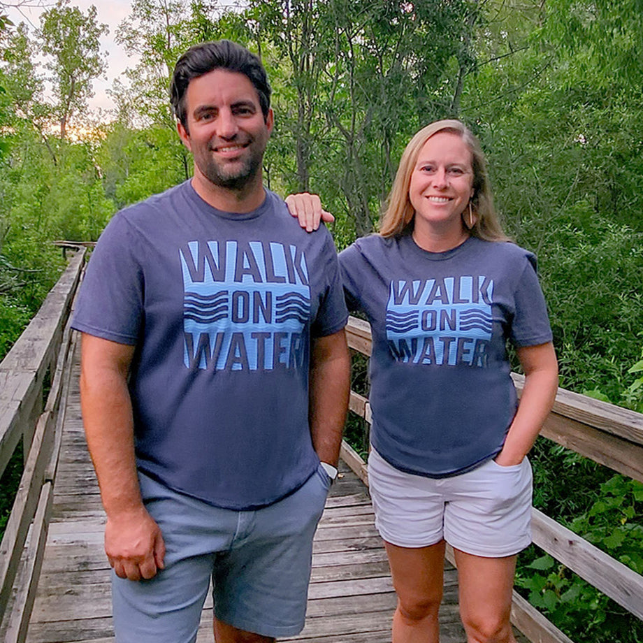 Husband and Wife wearing 'Walk on Water' Christian T Shirts standing on a wooden bridge with greenery in the background.